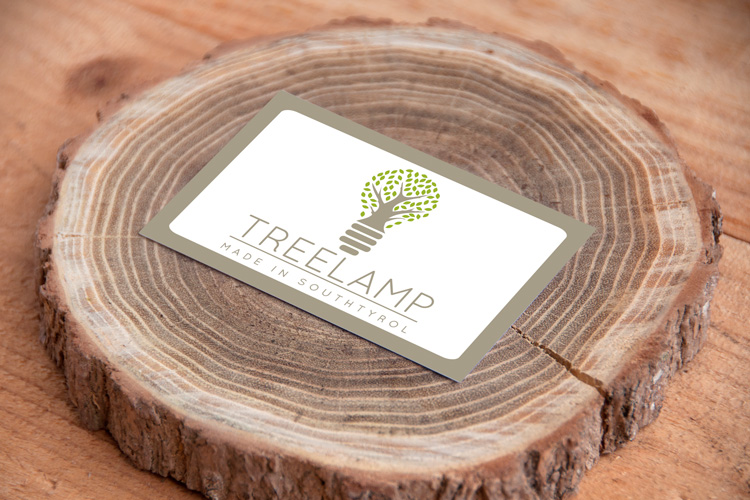 Realizzazione grafica biglietto da visita Treelamp formato rettangolare:logo centrale rappresentante una lampadina|albero con il naming e pay-off sottostante su fondo bianco inserito in una cornice con ancoli arrotondati verde|grigio, il tutto posizionato su un tronco d'albero tagliato